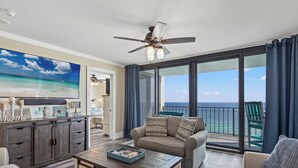 A spacious living room with a large flat-screen TV, a ceiling fan, and a view of the ocean through large windows. - Watercrest 1704 - Serenity Shores (Panama City Beach)