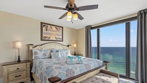 A bedroom with a large bed, a ceiling fan, a nightstand with a lamp, a bench, and a view of the ocean. - Watercrest 1704 - Serenity Shores (Panama City Beach)