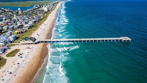 Une vue aérienne d’une plage avec une longue jetée qui s’avance dans l’océan, entourée de bâtiments résidentiels et sous un ciel dégagé.