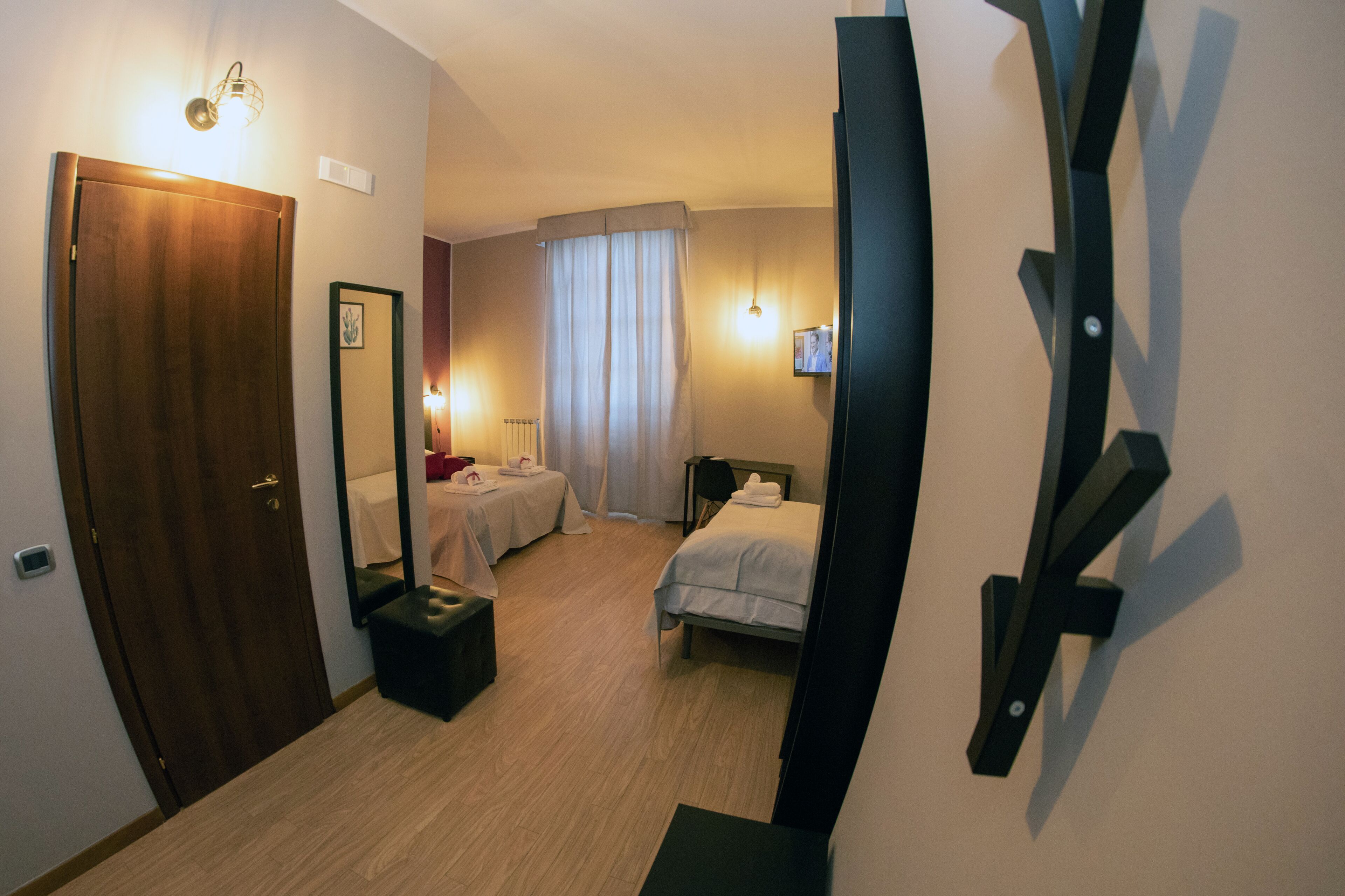 Habitación de hotel con puerta de madera, cama, mesita de noche, espejo, ventana con cortinas y televisor montado en la pared.