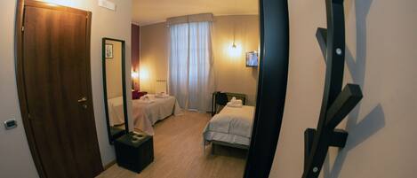 Habitación de hotel con puerta de madera, cama, mesita de noche, espejo, ventana con cortinas y televisor montado en la pared.