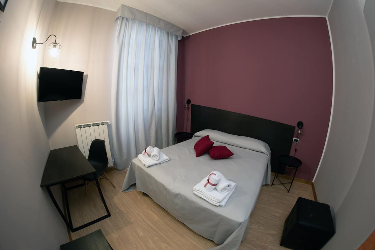 A neatly arranged hotel room with a bed, a small desk, a chair, a TV, and a bedside table.