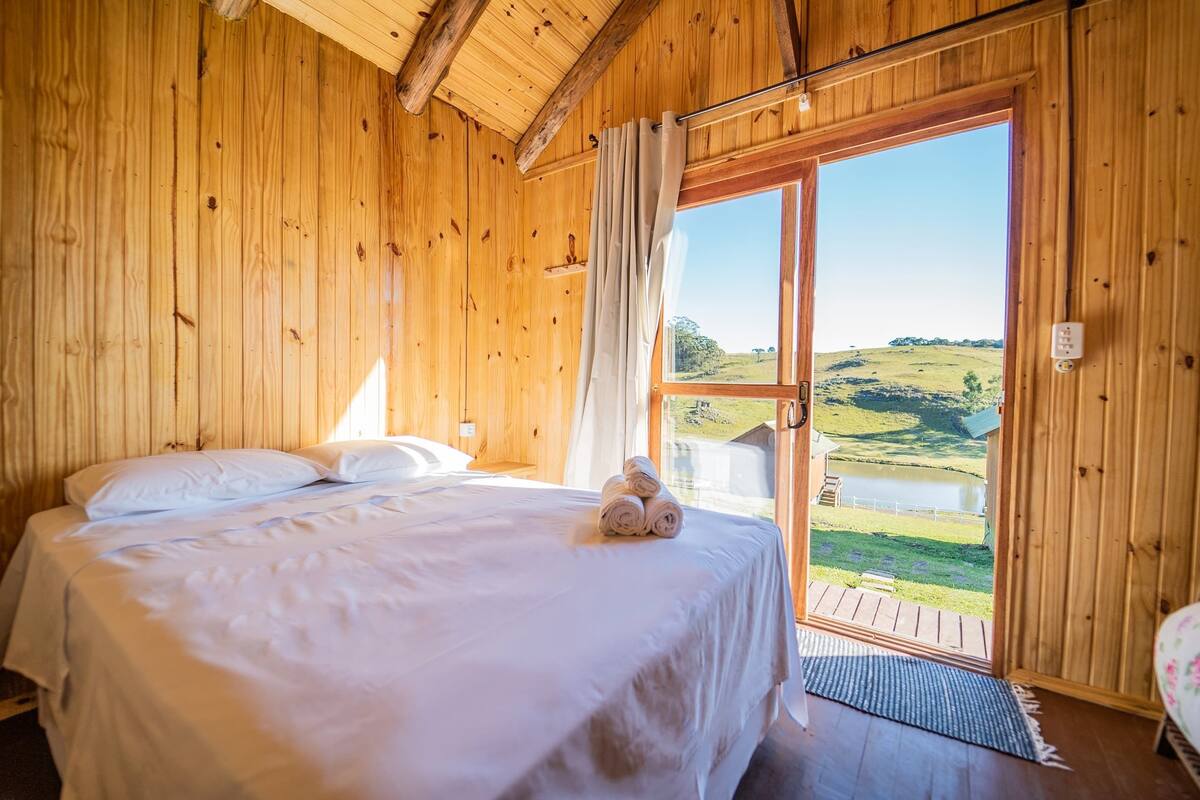 Een slaapkamer in een houten chalet met een bed, kussens en uitzicht op een meer en heuvels.