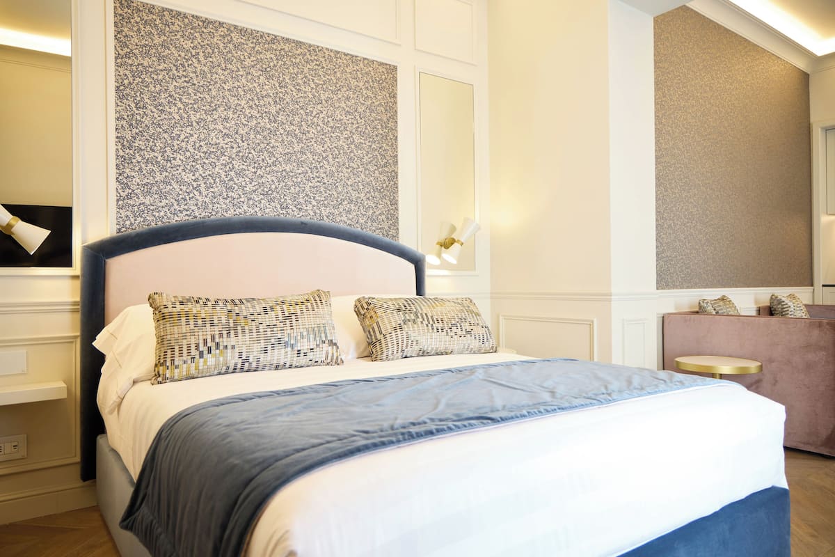 A neatly made bed with a blue and white bedspread, a patterned pillow, and a headboard. There is a small round table with a gold base and a white top. The room has a large mirror on the wall and a textured wall panel above the bed.