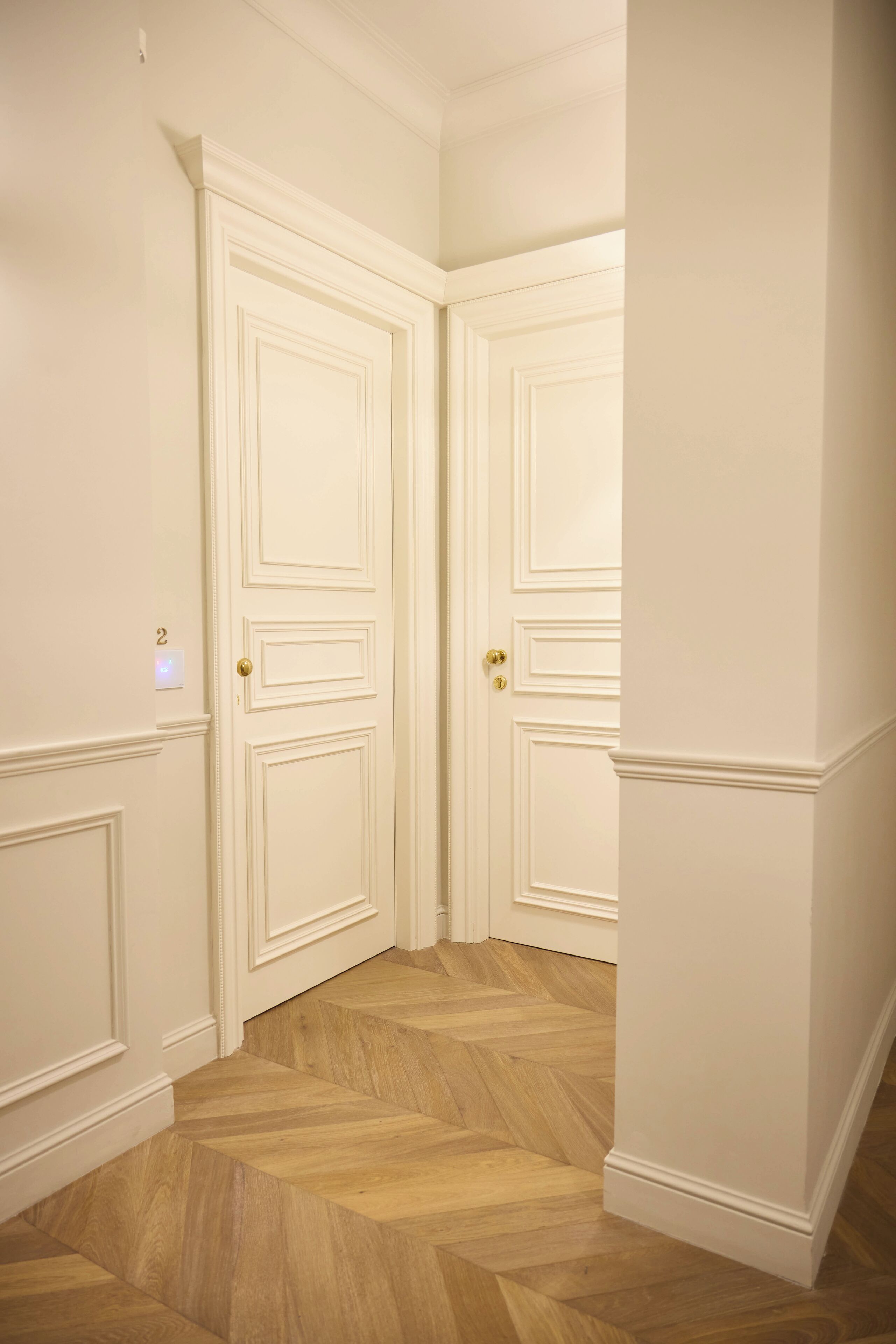 A hallway with two white doors, a light switch, and a herringbone-patterned wooden floor.