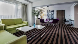 Une chambre d’hôtel moderne avec un canapé vert, une table basse, une moquette à rayures, un coin repas avec des chaises et un bureau.