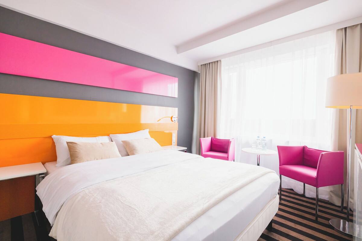 Une chambre d’hôtel avec un grand lit, une petite table, deux chaises et une fenêtre avec des rideaux.