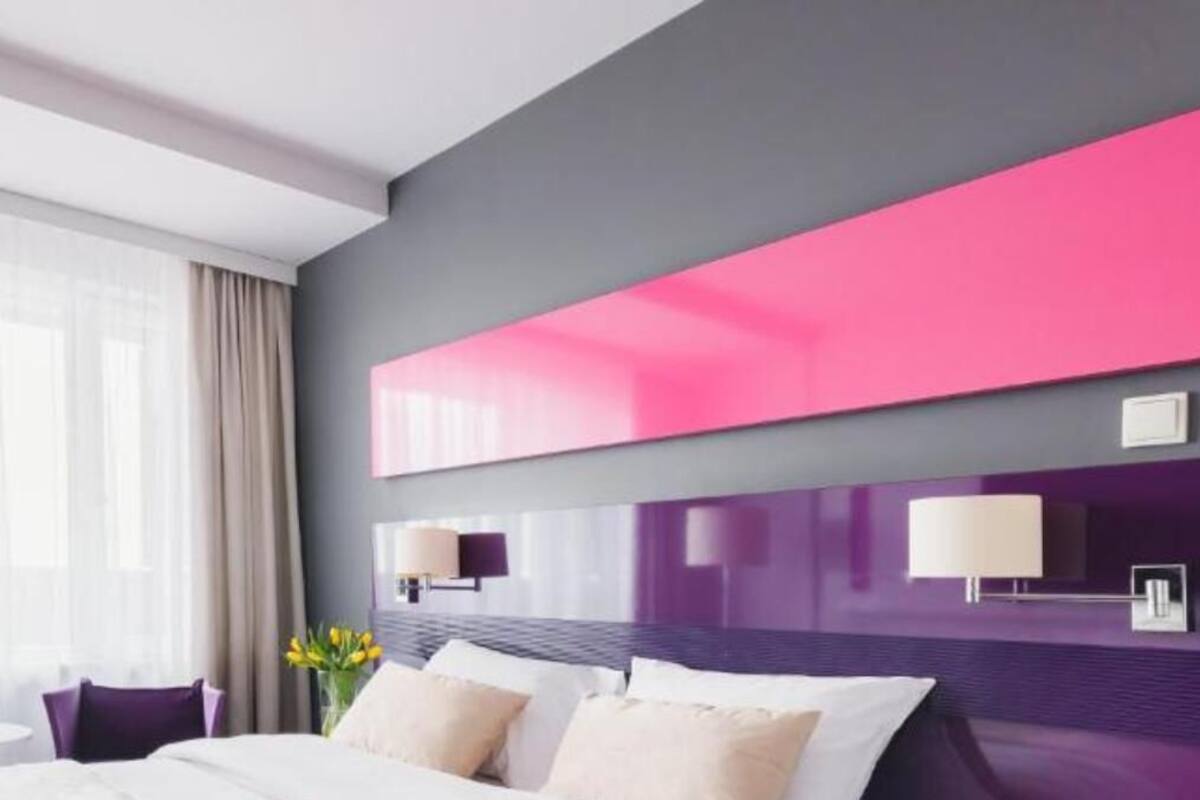 Une chambre d’hôtel moderne avec un grand lit, une petite table sur laquelle se trouve une bouteille d’eau, un vase avec des fleurs et un grand panneau mural aux couleurs rose et violet.