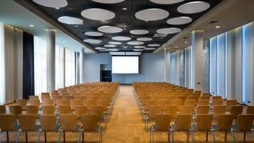 Un salón de conferencias con filas de sillas de madera, una pantalla grande al frente y luces circulares en el techo.