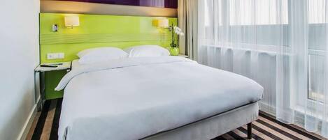 Habitación de hotel con una cama grande, cabecera verde, mesita de noche, lámpara y ventana con cortinas translúcidas.