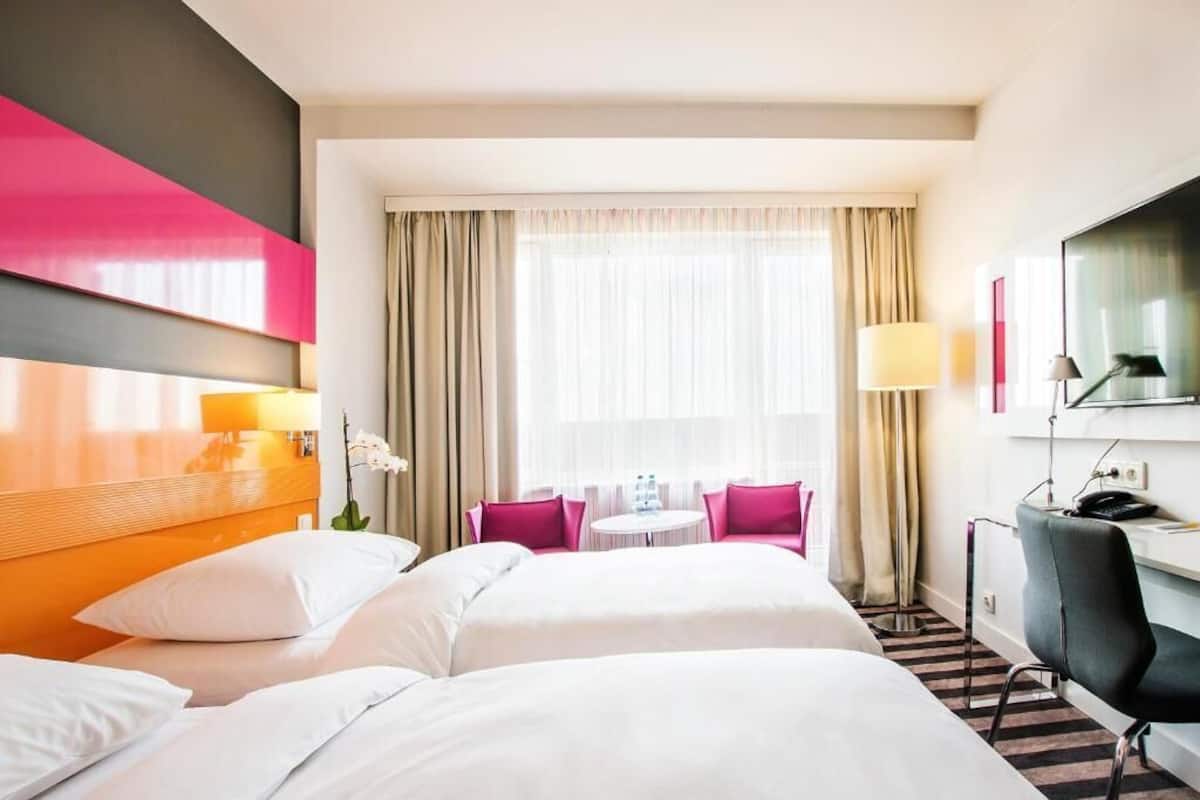 Une chambre d’hôtel avec un grand lit, un bureau avec une chaise, une petite table avec des chaises et une grande fenêtre avec des rideaux.