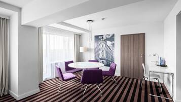 A hotel room with a round table, purple chairs, a glass desk, a large window with curtains, and a striped carpet.