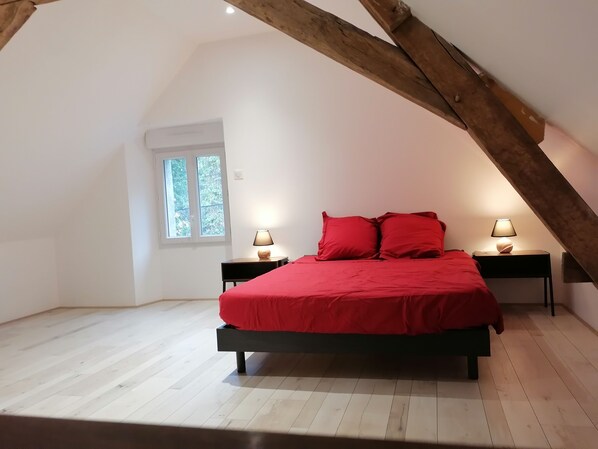 Ein Schlafzimmer mit schrägem Dach, einem Bett mit roter Bettwäsche, zwei Nachttischen mit Lampen und einem Fenster mit Blick auf Bäume.