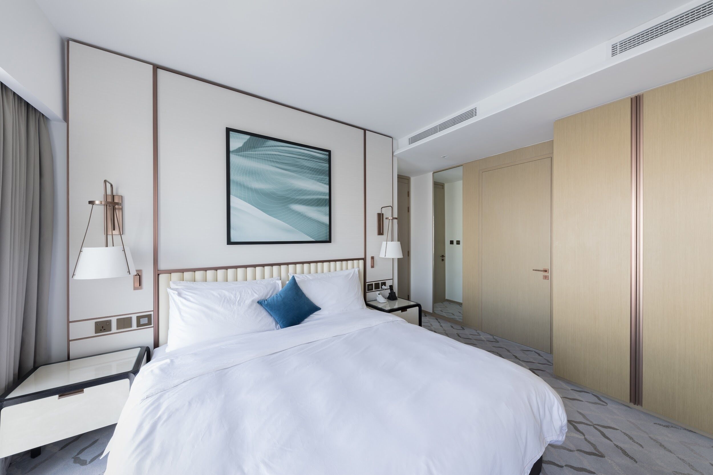 A hotel room with a large bed, a nightstand, a bedside table, a lamp, a wall-mounted artwork, and a wardrobe.