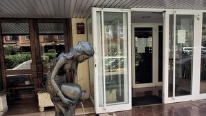 Une entrée d’immeuble ornée d’une sculpture métallique représentant une figure versant de l’eau, une porte d’entrée en verre et une enseigne sur laquelle on peut lire « FUEGO ».