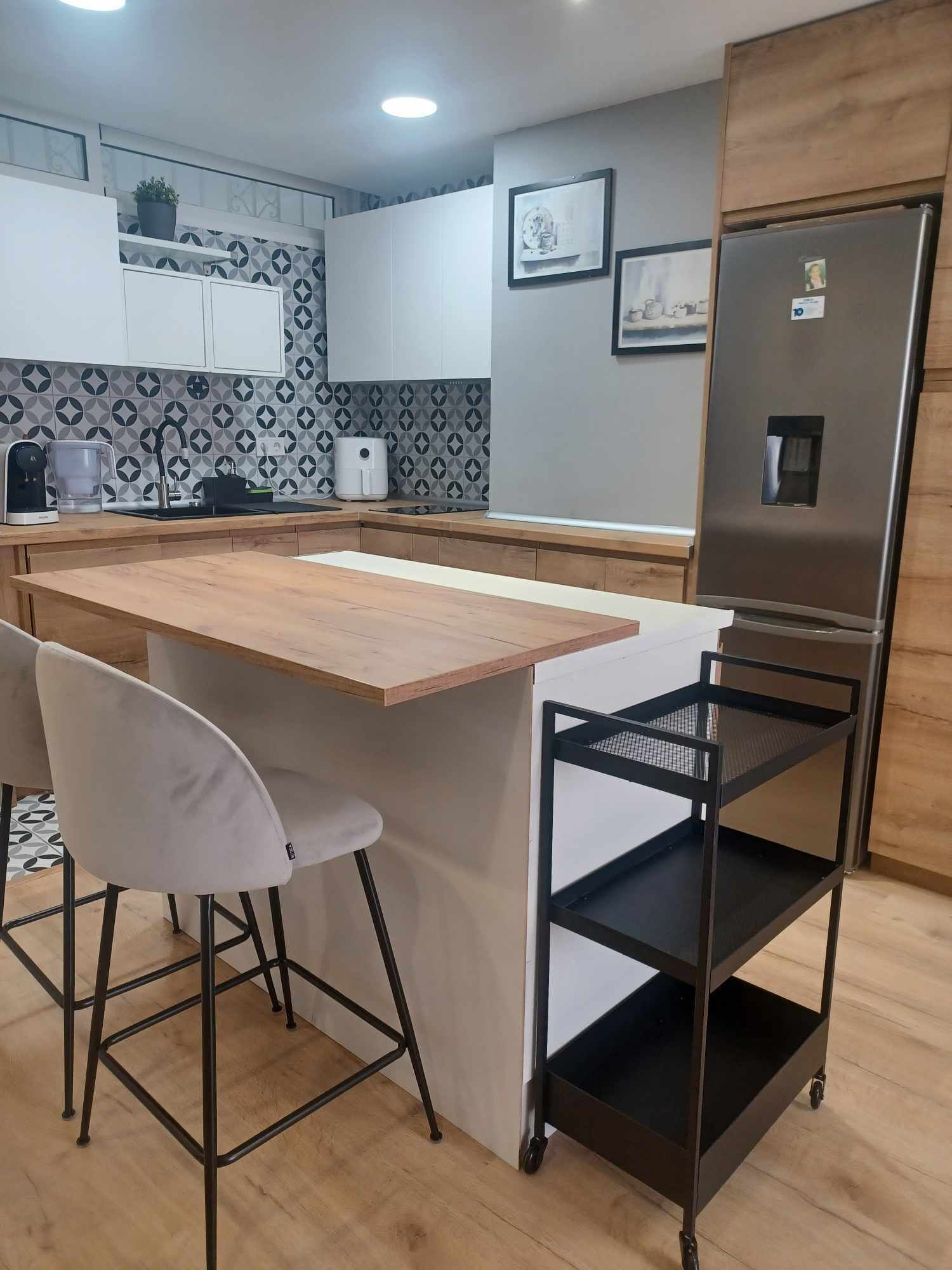 A modern kitchen with a wooden countertop, two white bar stools, a stainless steel refrigerator, and a patterned backsplash.