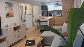 Un salon moderne avec un canapé, une table basse et un téléviseur. On aperçoit au fond une cuisine équipée d’un four à micro-ondes et de placards.