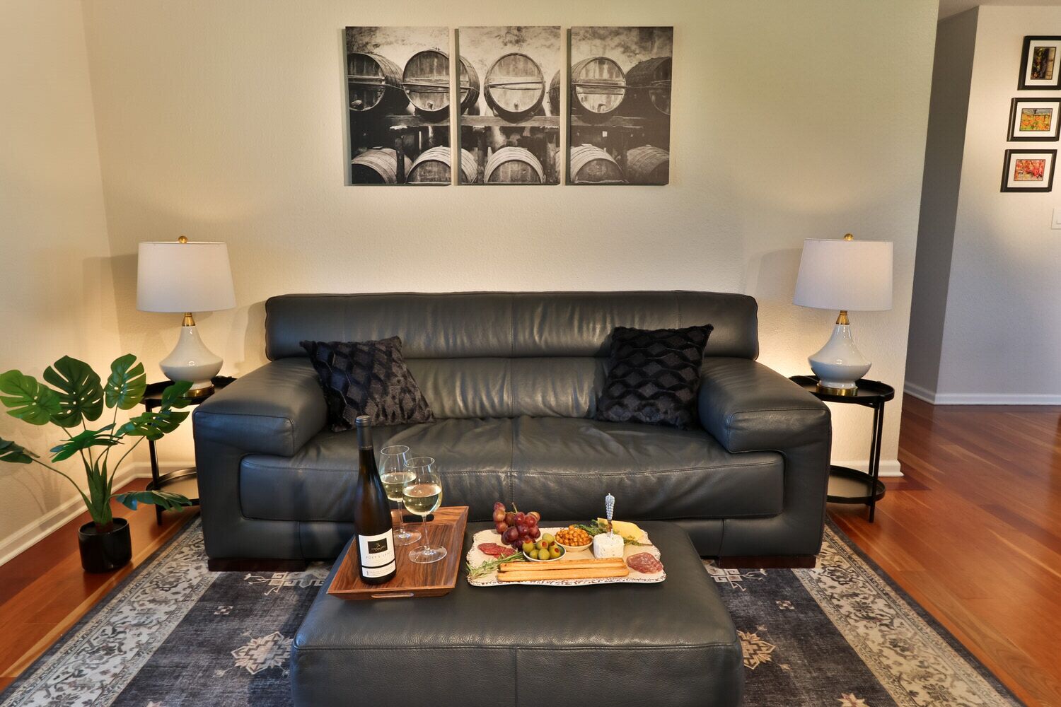A modern living room with a black leather sofa, two side tables with lamps, a coffee table with a wine bottle and glasses, a tray with cheese and fruits, and a large abstract painting on the wall.