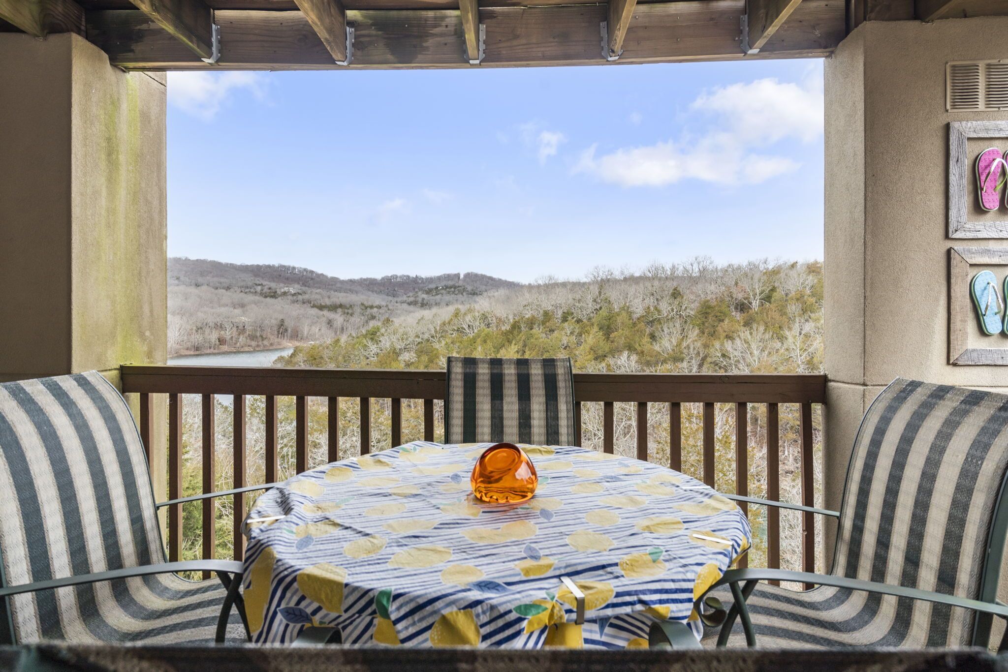 A round table with a striped tablecloth and a single orange glass on it, surrounded by striped chairs, with a view of a hilly landscape through a wooden pergola.