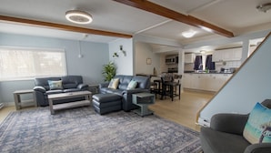Un salon moderne avec un canapé d’angle, une table basse et un tapis à motifs. On y trouve également un coin repas avec une table et des chaises, et une cuisine en arrière-plan.