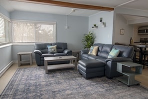 A living room with a sofa, a coffee table, and a rug. There is a window with blinds, a plant, and a kitchen area in the background.