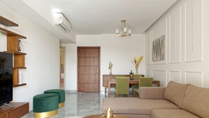 A modern living room with a sectional sofa, a coffee table, and a TV on a wooden stand. There is a dining area with a table and chairs, and a wooden door in the background. - VESTA - New Cairo Residence (New Cairo)