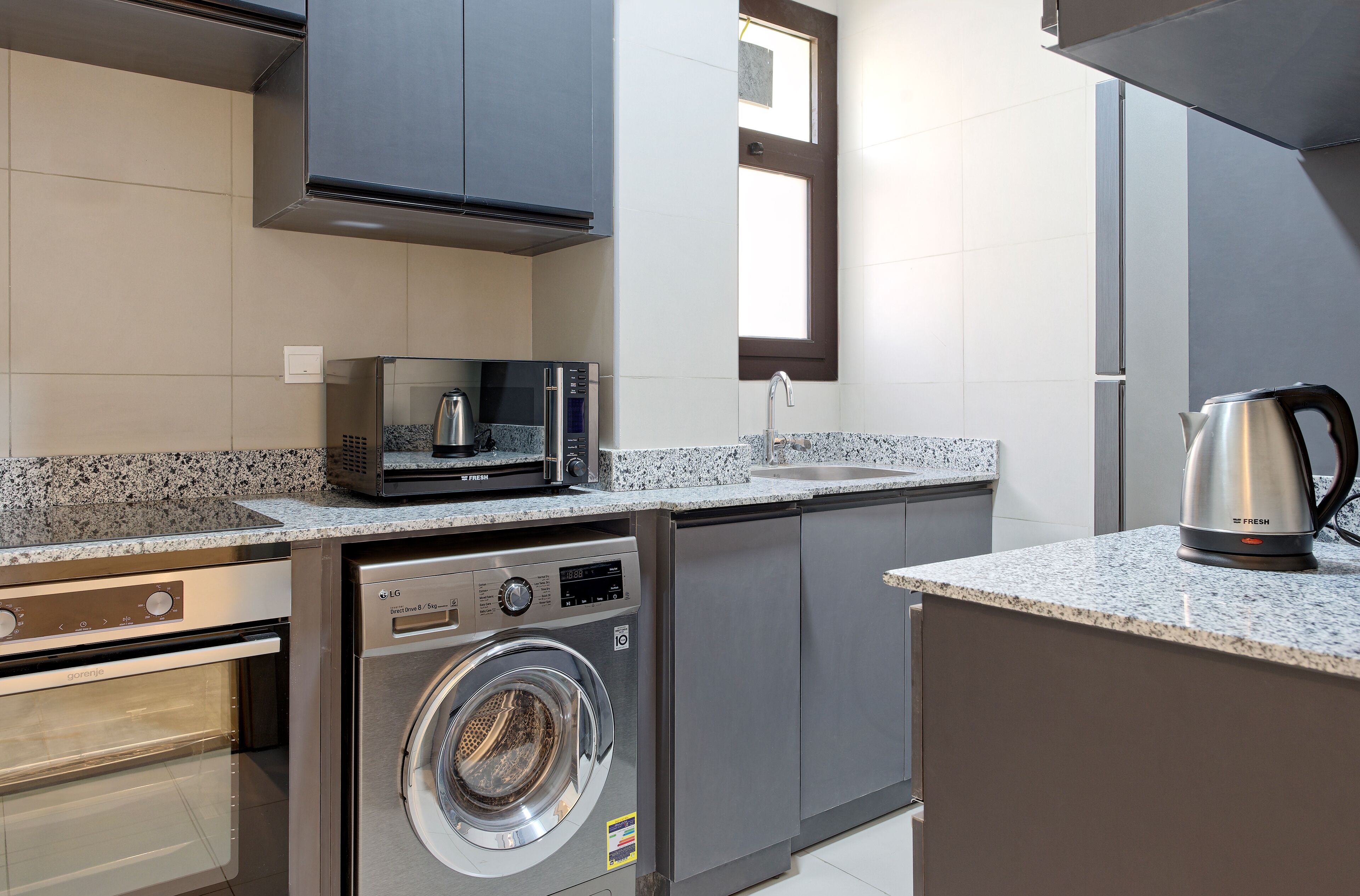 Cocina compacta con lavadora y secadora integradas, microondas y hervidor sobre la encimera.