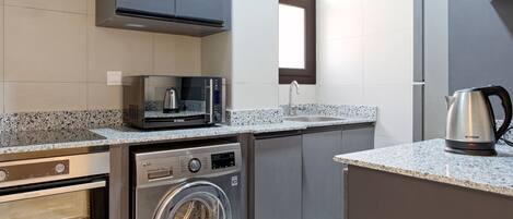 Cocina compacta con lavadora y secadora integradas, microondas y hervidor sobre la encimera.
