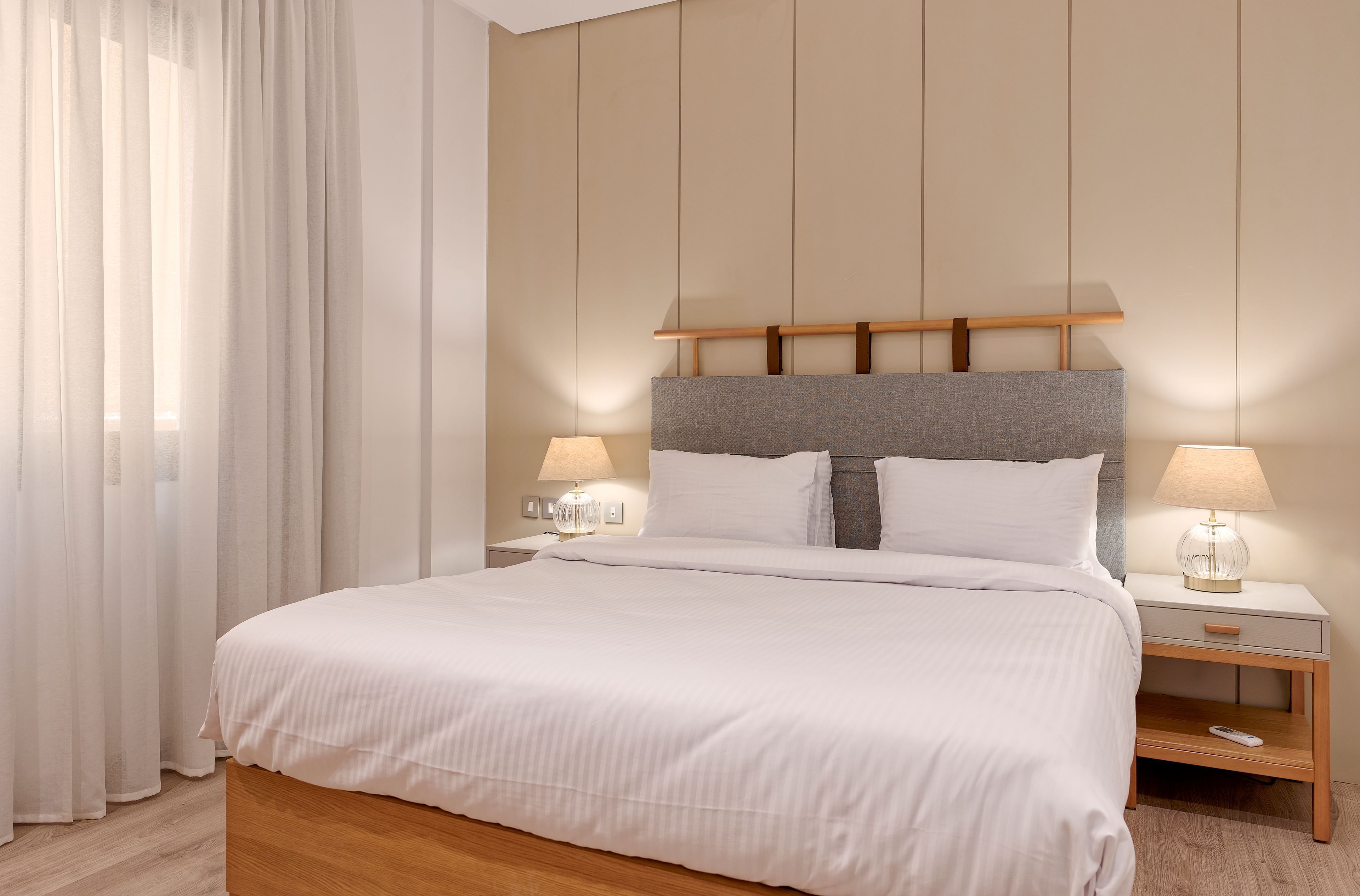 A neatly made bed with white linens, a wooden headboard, two bedside tables with lamps, and a large window with sheer curtains.