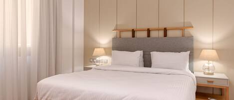 A neatly made bed with white linens, a wooden headboard, two bedside tables with lamps, and a large window with sheer curtains.