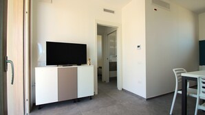 A modern, minimalistic room with a flat-screen TV on a white cabinet, a dining table with chairs, and a visible door leading to another room. - Athesis Lodges (Rosolina Mare)