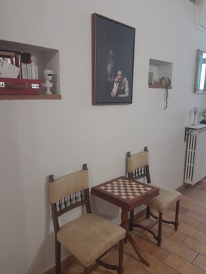 A room with two wooden chairs, a chess table, a framed painting, and a small shelf with books and a bust.