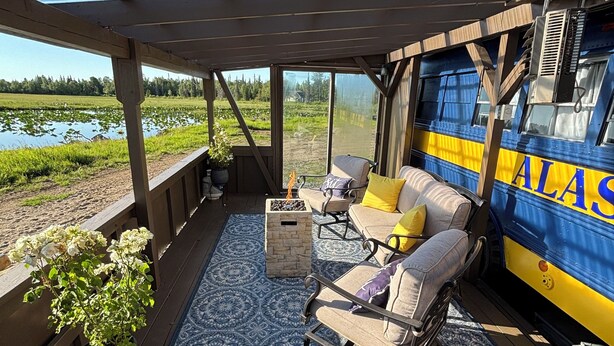 Un espace extérieur aménagé avec des sièges, abrité par un toit, comprenant un foyer, un tapis à motifs bleus et offrant une vue sur un lac et une forêt.