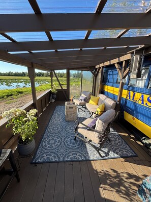 A covered outdoor seating area with a fire pit, a blue patterned rug, and a view of a lake and forest.