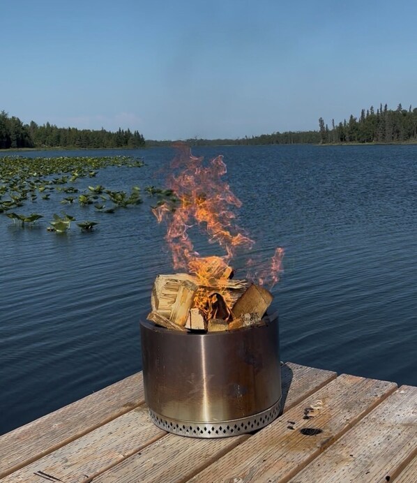 A metal fire pit with burning wood on a wooden dock by a lake.