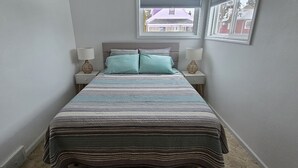 Cama bem arrumada com roupa de cama listrada, duas luminárias de cabeceira e uma janela com vista para prédios.