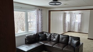 Um quarto com um sofá seccional de couro marrom escuro, uma janela grande com cortinas florais e piso coberto de carpete.