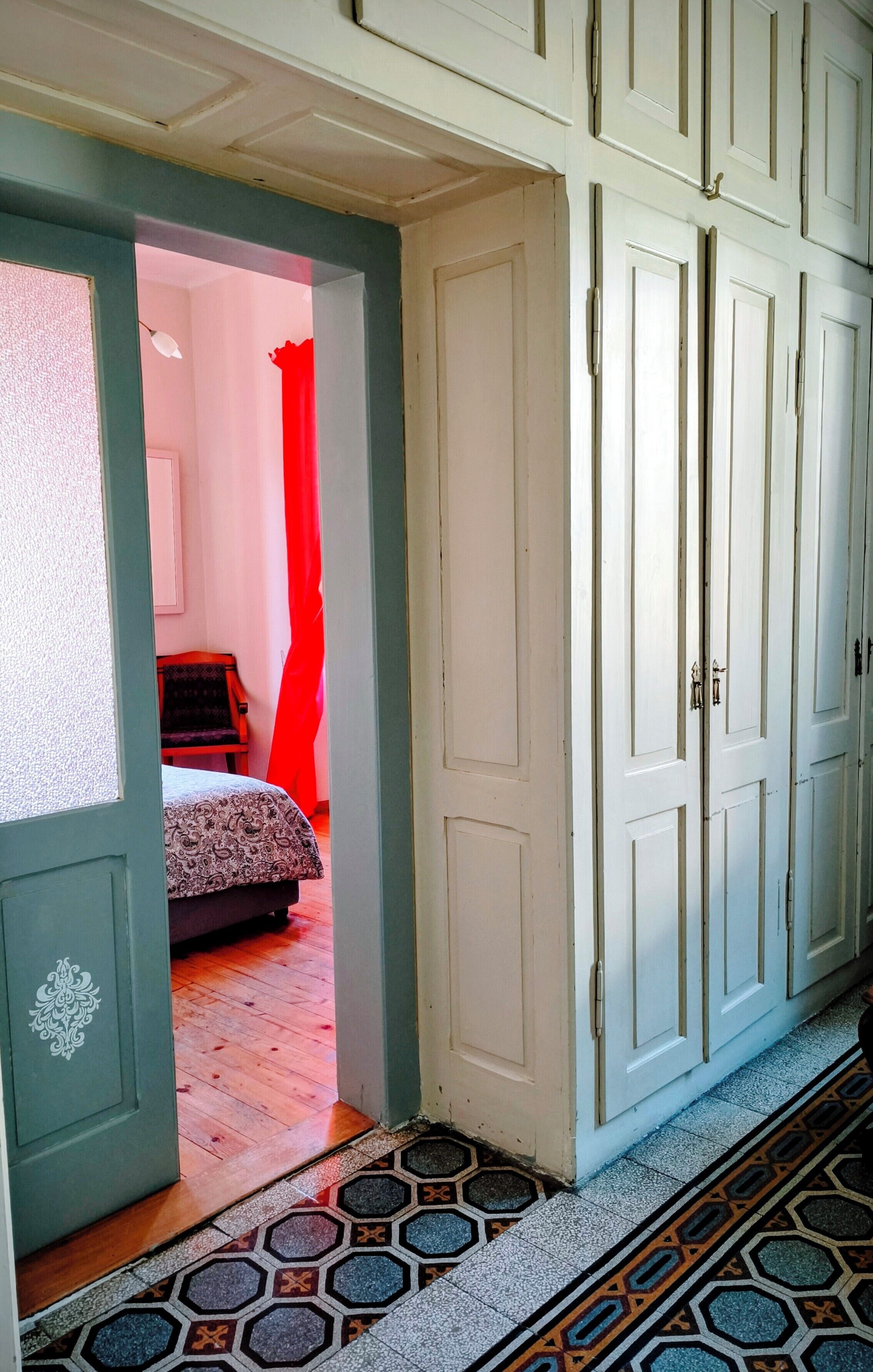 Ein Flur mit einem gemusterten Boden, einem Bett mit einer gemusterten Tagesdecke und einem roten Vorhang in einem Zimmer.