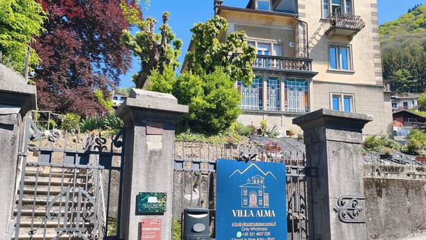 Una vila amb entrada tancada per reixa, un cartelló blau i un edifici amb balcons.