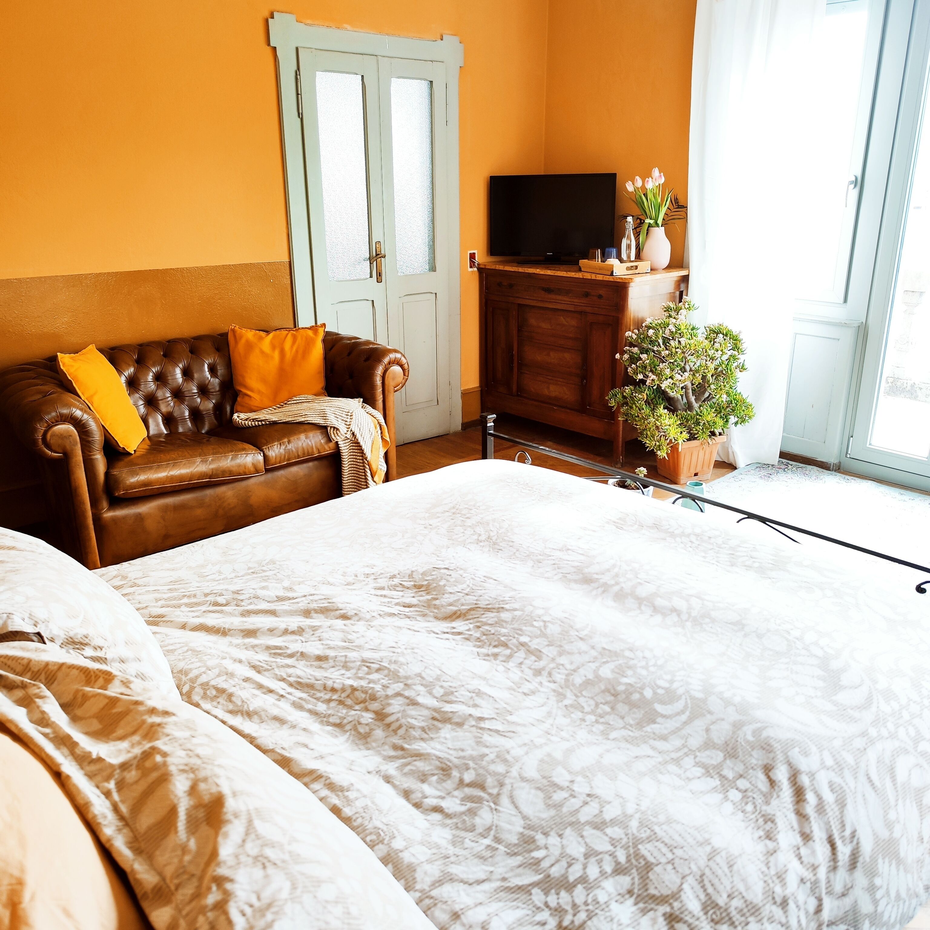 A room with a brown leather sofa, a white patterned bedspread, a wooden cabinet with a flat-screen TV, and a potted plant.