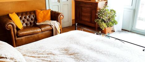 A room with a brown leather sofa, a white patterned bedspread, a wooden cabinet with a flat-screen TV, and a potted plant.
