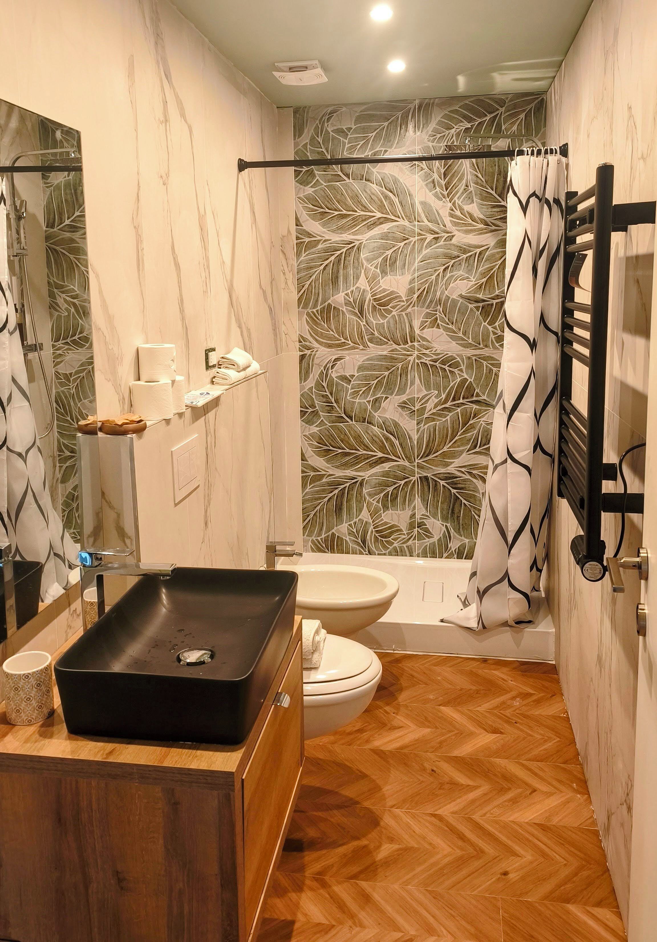 Un baño con suelo de madera, un lavabo negro, inodoro, ducha con pared estampada con hojas y un perchero para toallas.