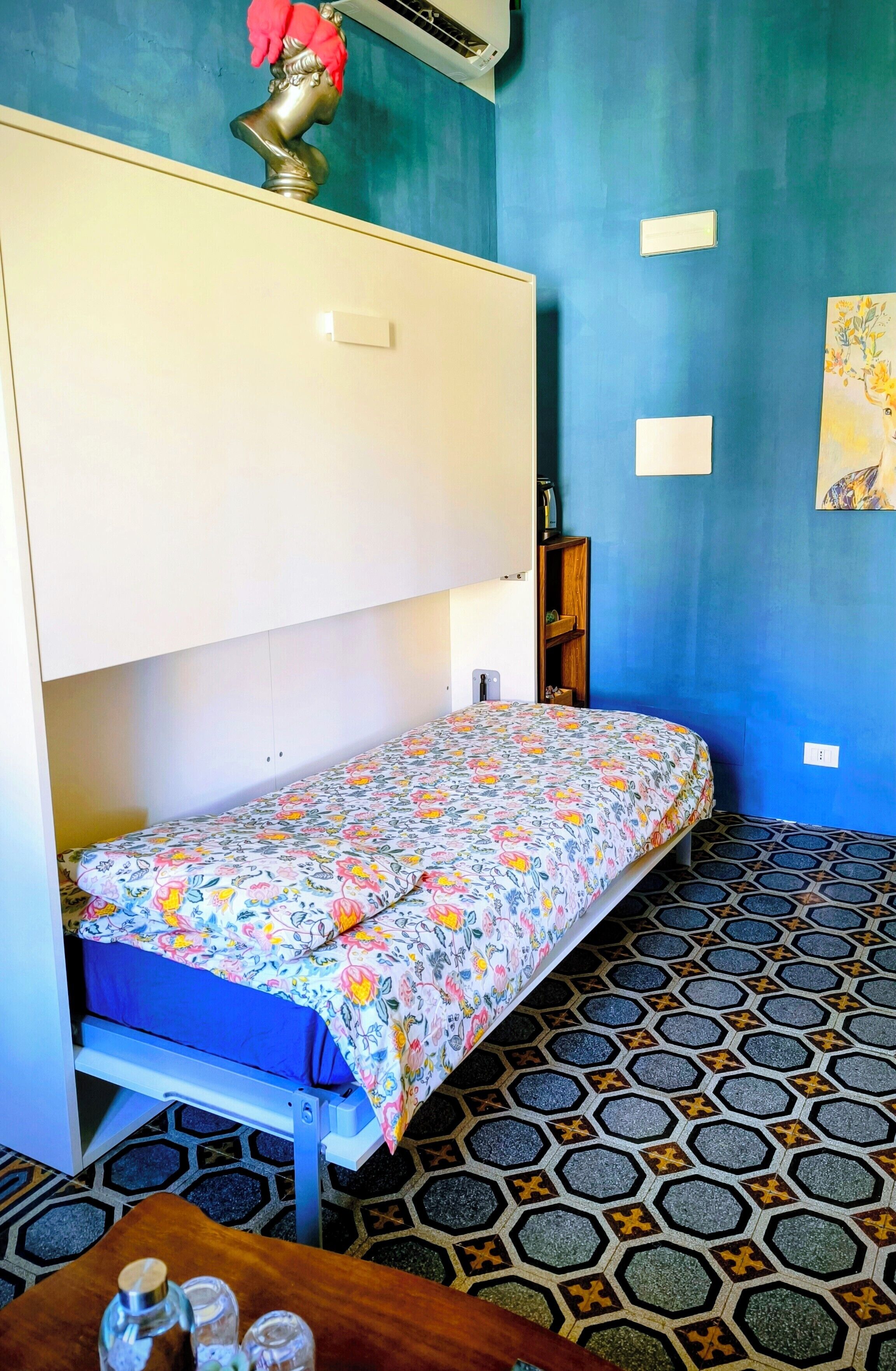 A room with a bed, a small table with a bottle and glass, a shelf with items, and a wall-mounted air conditioner.