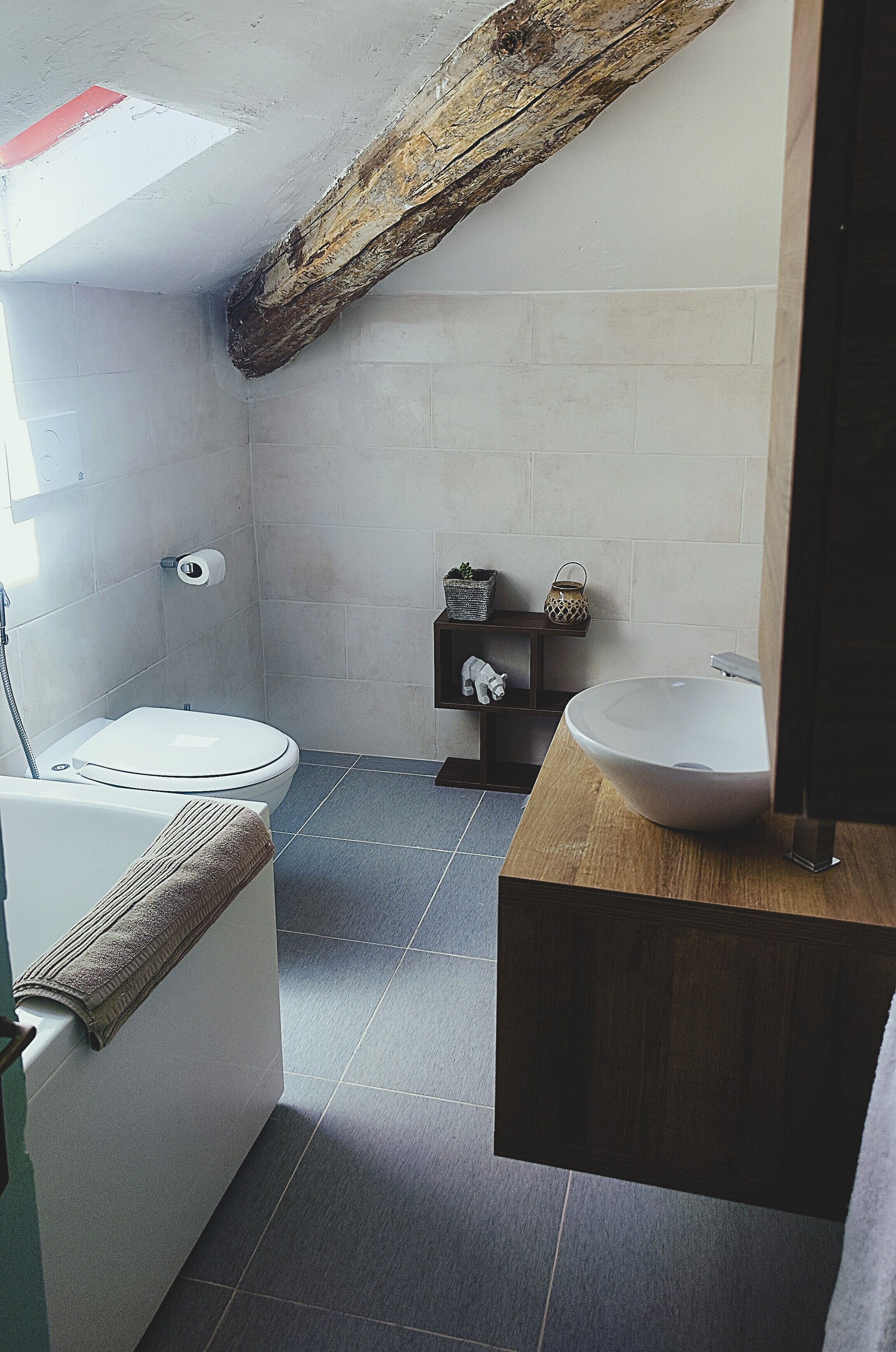 Une salle de bain avec des toilettes blanches, un meuble-lavabo en bois doté d’un grand évier et une étagère ornée d’objets décoratifs.