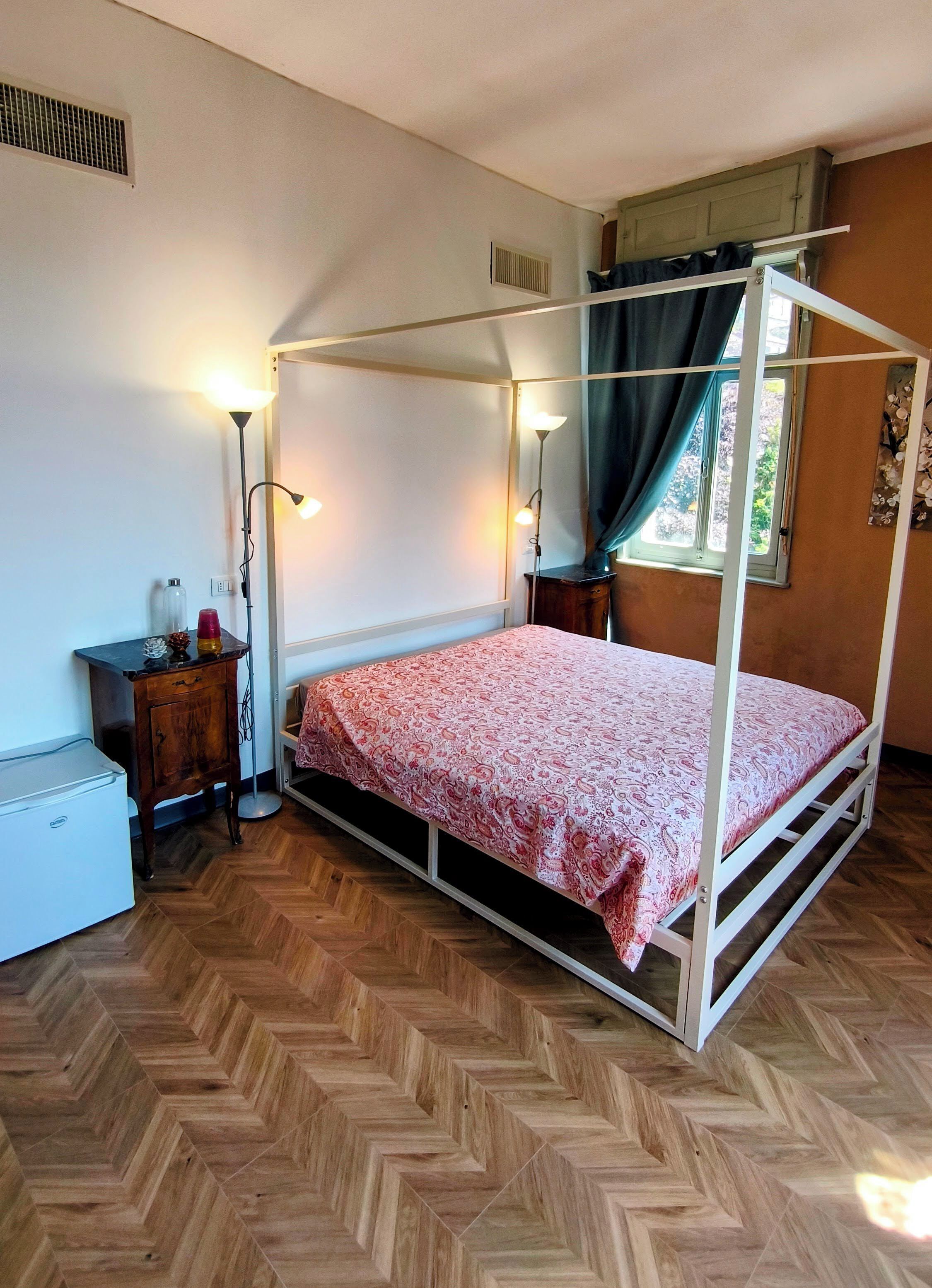 Ein Zimmer mit einem Einzelbett, einem Holzboden mit Fischgrätmuster, einem Nachttisch mit Lampe, einem kleinen Kühlschrank und einem Fenster mit Vorhängen.