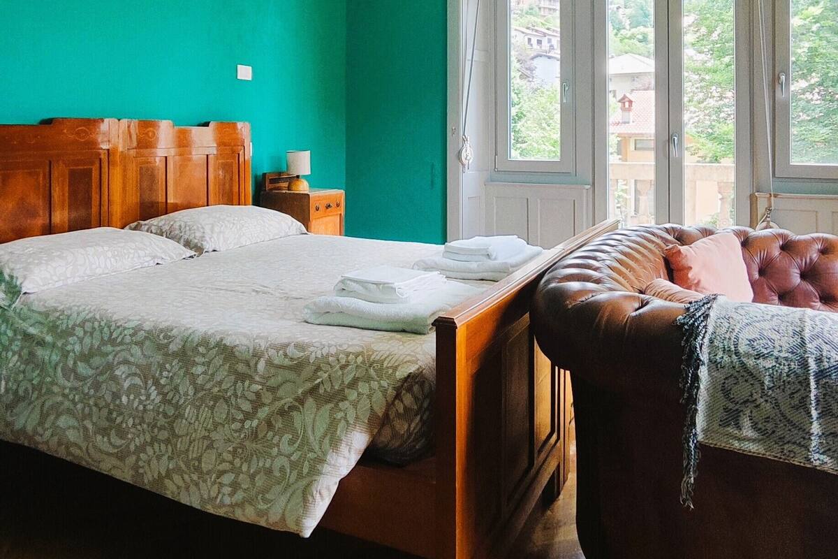 A bedroom with a wooden bed, a wooden nightstand, a window with a view of greenery, and a brown leather chair.