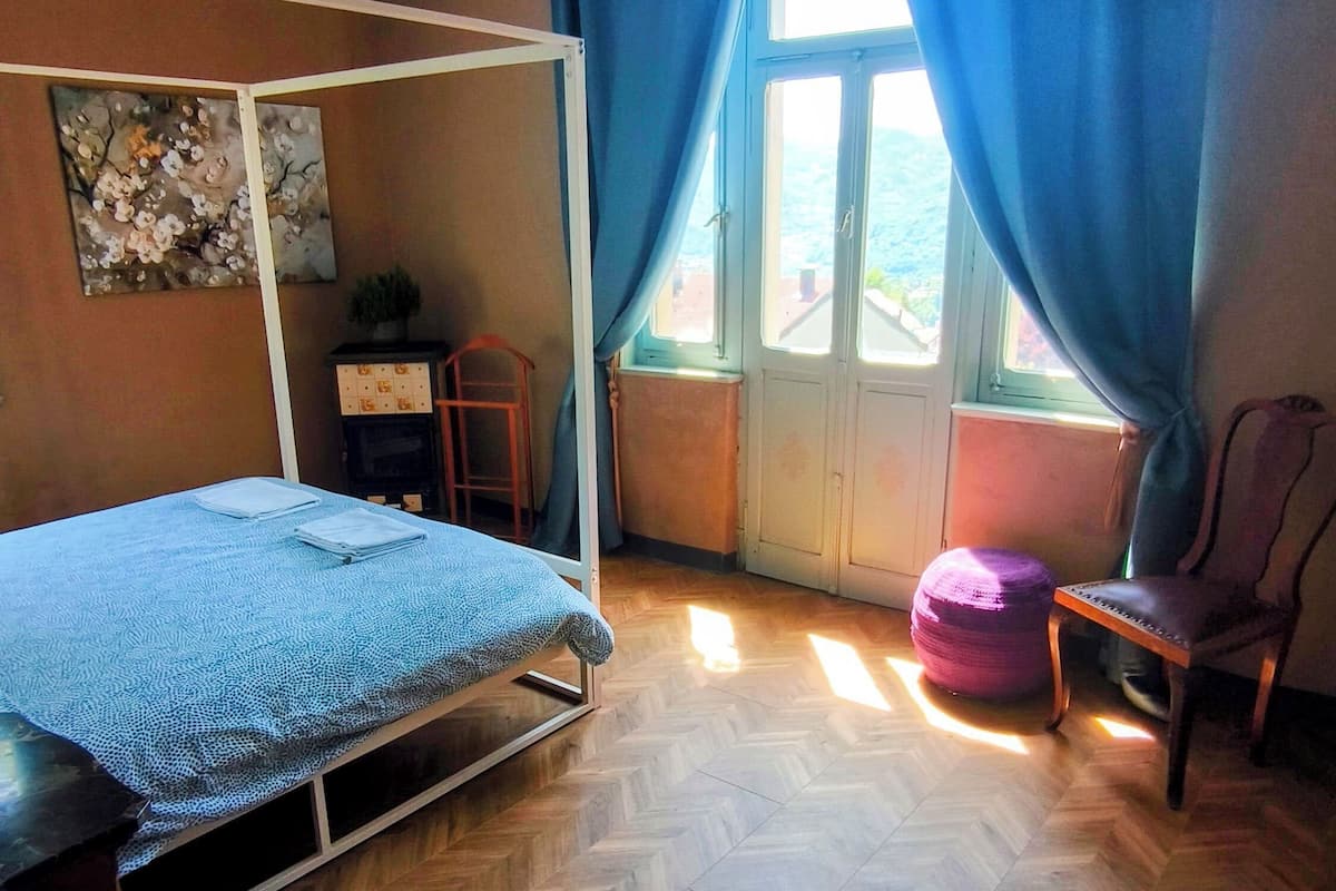 A room with a bed, a desk, a chair, a window with curtains, and a herringbone-patterned floor.