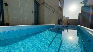 A swimming pool with blue mosaic tiles, a metal ladder, and a building with beige tiled walls and a window.