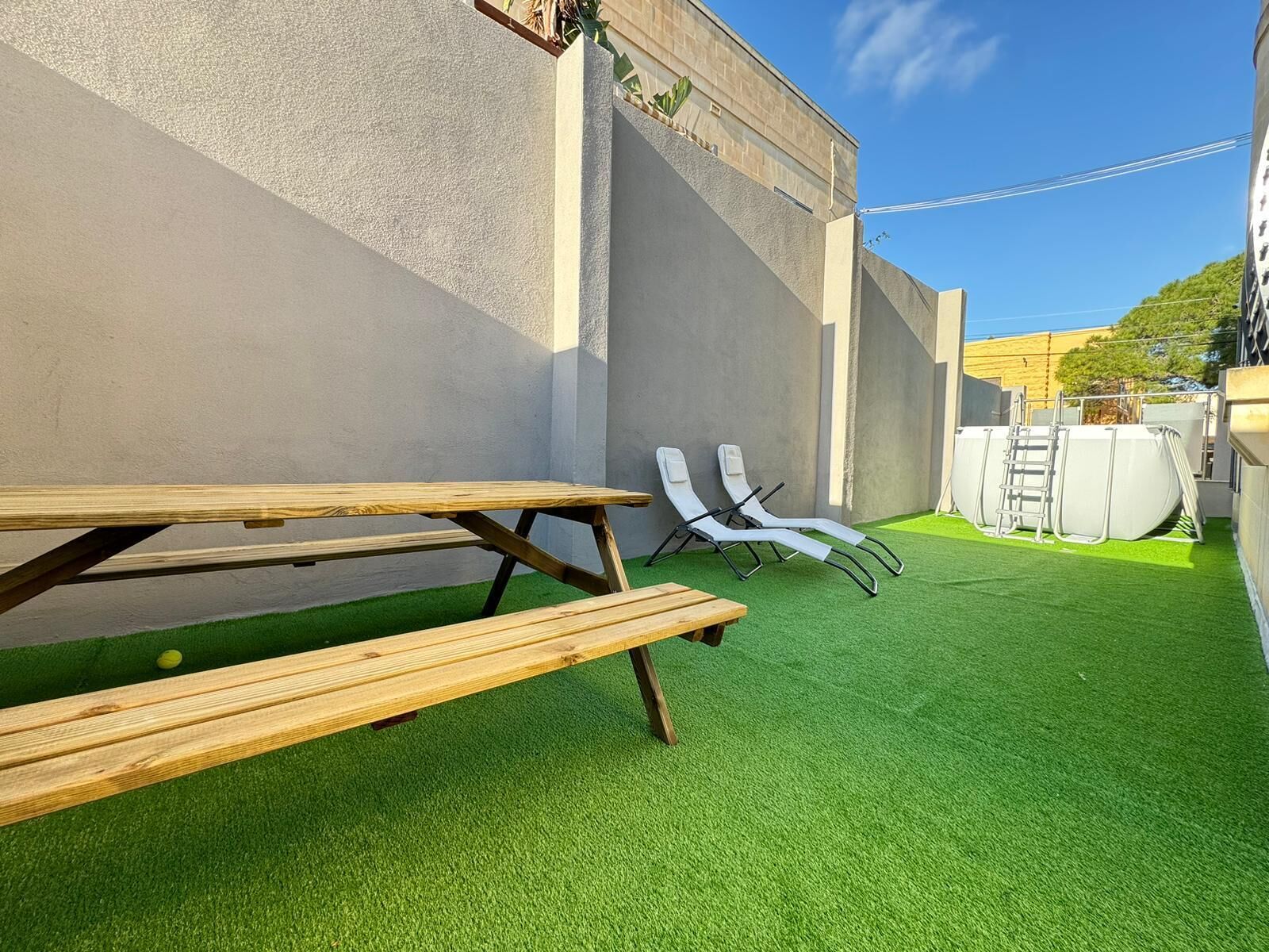 Un patio con una mesa de madera y dos sillones de descanso, rodeado por un muro bajo y césped artificial verde.