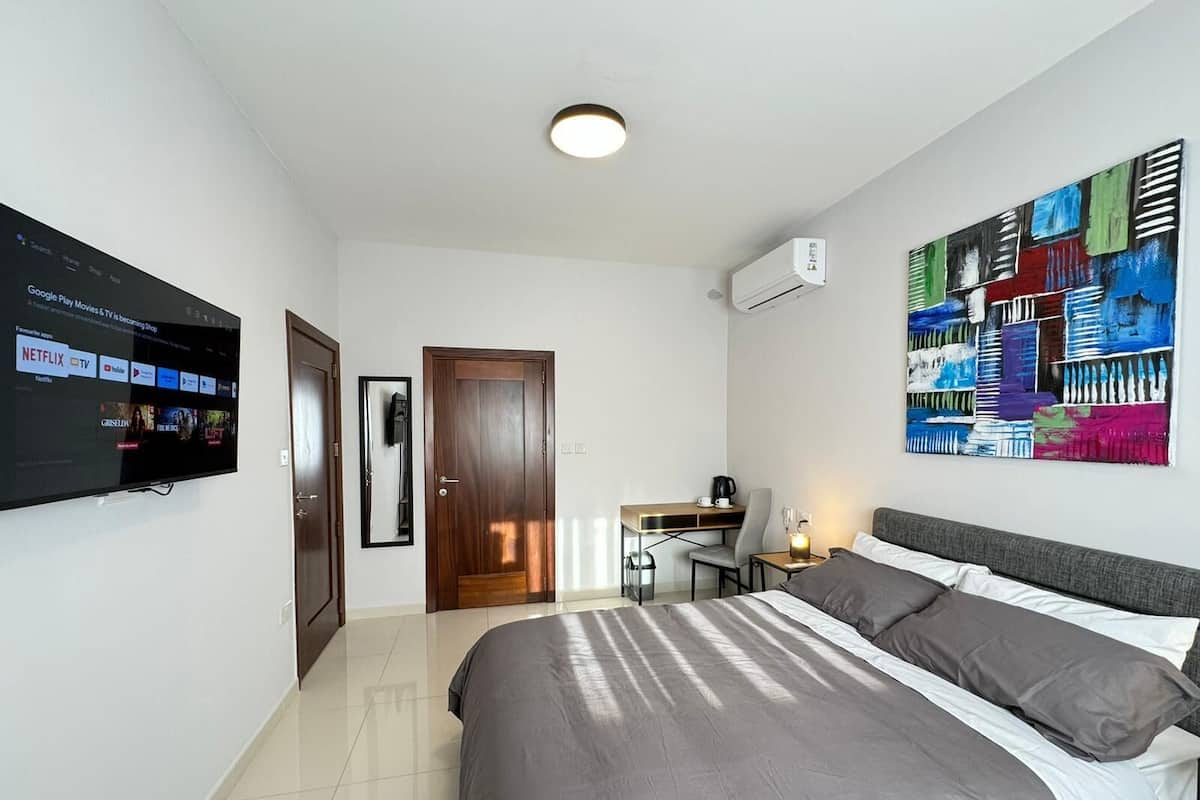 Ein modernes Hotelzimmer mit einem großen Bett, einem Schreibtisch mit Stuhl, einem flachbildfernseher an der Wand und einem farbenfrohen abstrakten Gemälde.